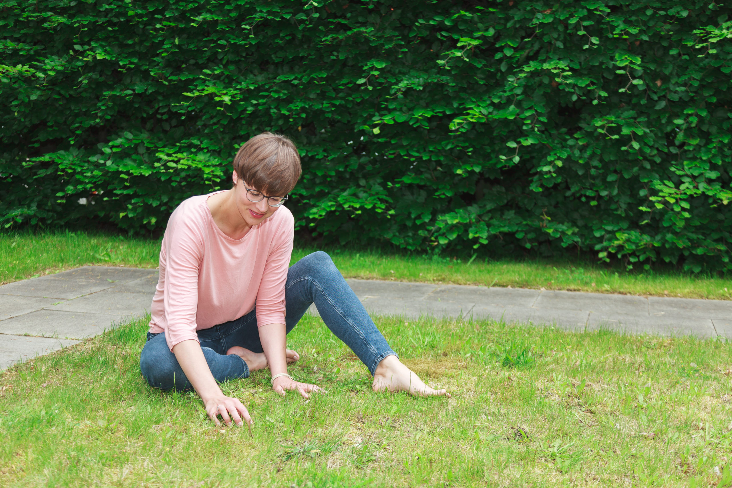 Eine Person mit Brille, rosa Hemd und Jeans sitzt barfuß im Gras, blickt nach unten und berührt den Boden, im Hintergrund eine grüne Hecke.