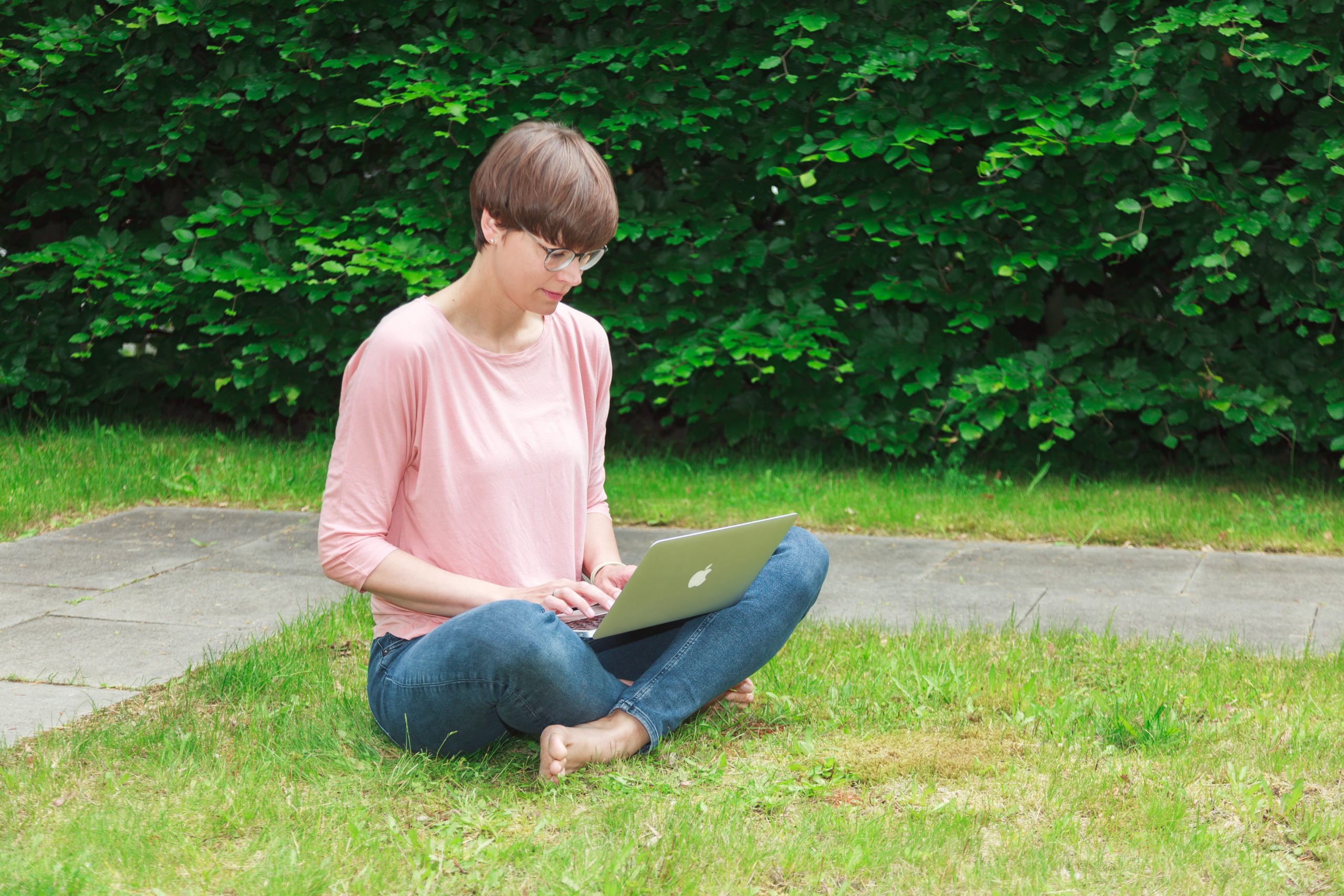 Eine Person sitzt im Schneidersitz im Gras und benutzt einen Laptop im Freien, in der Nähe eines gepflasterten Weges und dichter grüner Sträucher im Hintergrund.