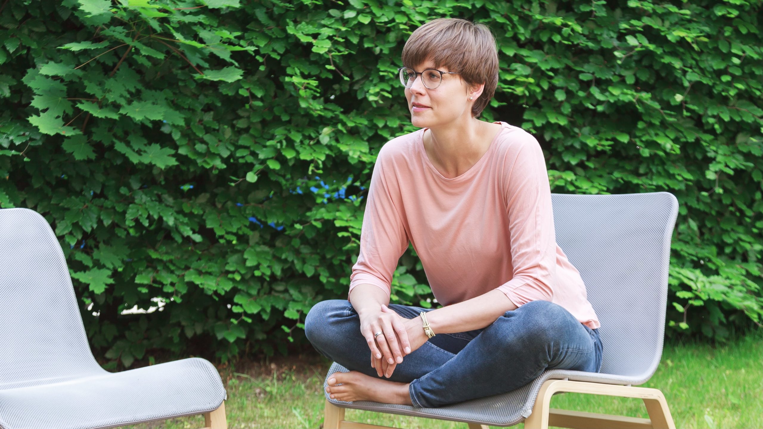 Eine Person mit kurzen Haaren und Brille sitzt im Schneidersitz auf einem Stuhl im Freien. Sie trägt ein rosa Oberteil und Jeans, im Hintergrund ist grünes Laub zu sehen.