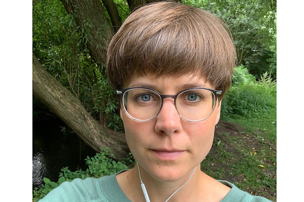 Eine Person mit kurzen braunen Haaren, Brille und Kopfhörern steht im Freien in einem grünen Waldgebiet.