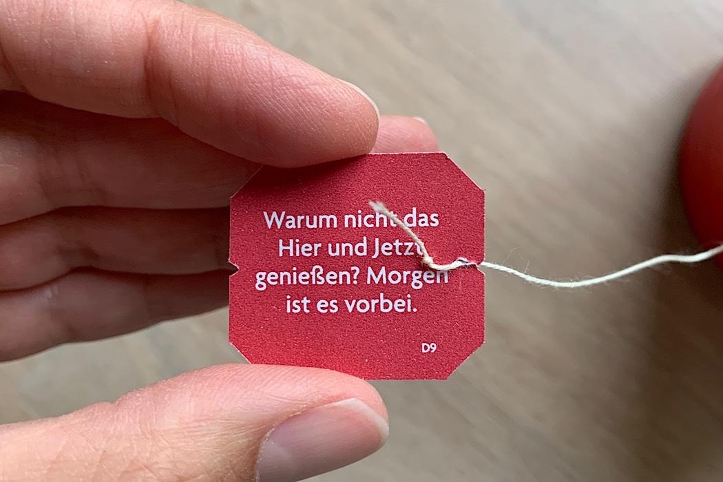 Eine Hand hält ein rotes Teebeutel-Etikett mit dem deutschen Text: "Warum nicht das Hier und Jetzt genießen? Morgen ist es vorbei." Eine weiße Schnur ist an dem Anhänger befestigt.