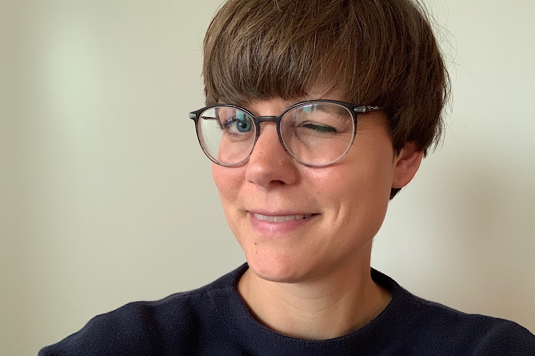 Person mit kurzen braunen Haaren und Brille, die einen dunklen Pullover trägt, lächelt und zwinkert in die Kamera vor einem schlichten hellen Hintergrund.
