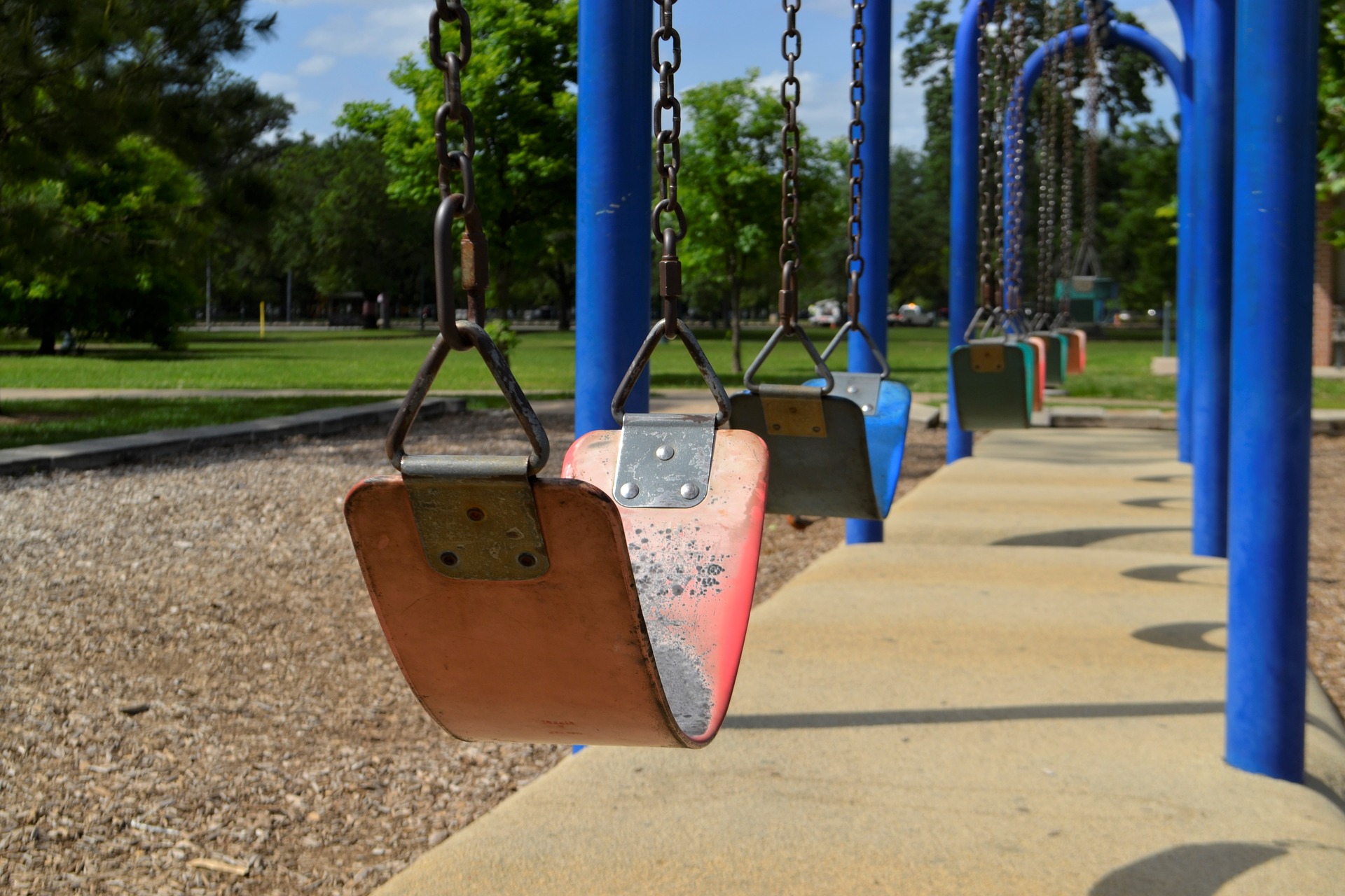 Leere Schaukeln hängen an blauen Gestellen in einer Reihe auf einem Spielplatz an einem sonnigen Tag, mit Bäumen und einer grasbewachsenen Parkfläche im Hintergrund.