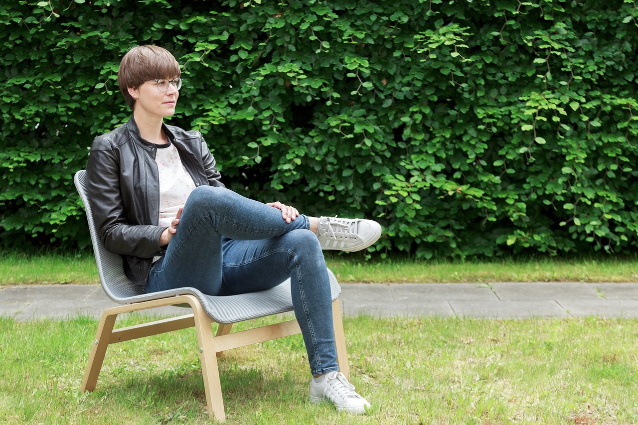 Eine Person mit kurzen Haaren, Brille, Jeans und schwarzer Jacke sitzt auf einem Stuhl mit gekreuzten Beinen auf einer Wiese, mit einer grünen Hecke im Hintergrund.