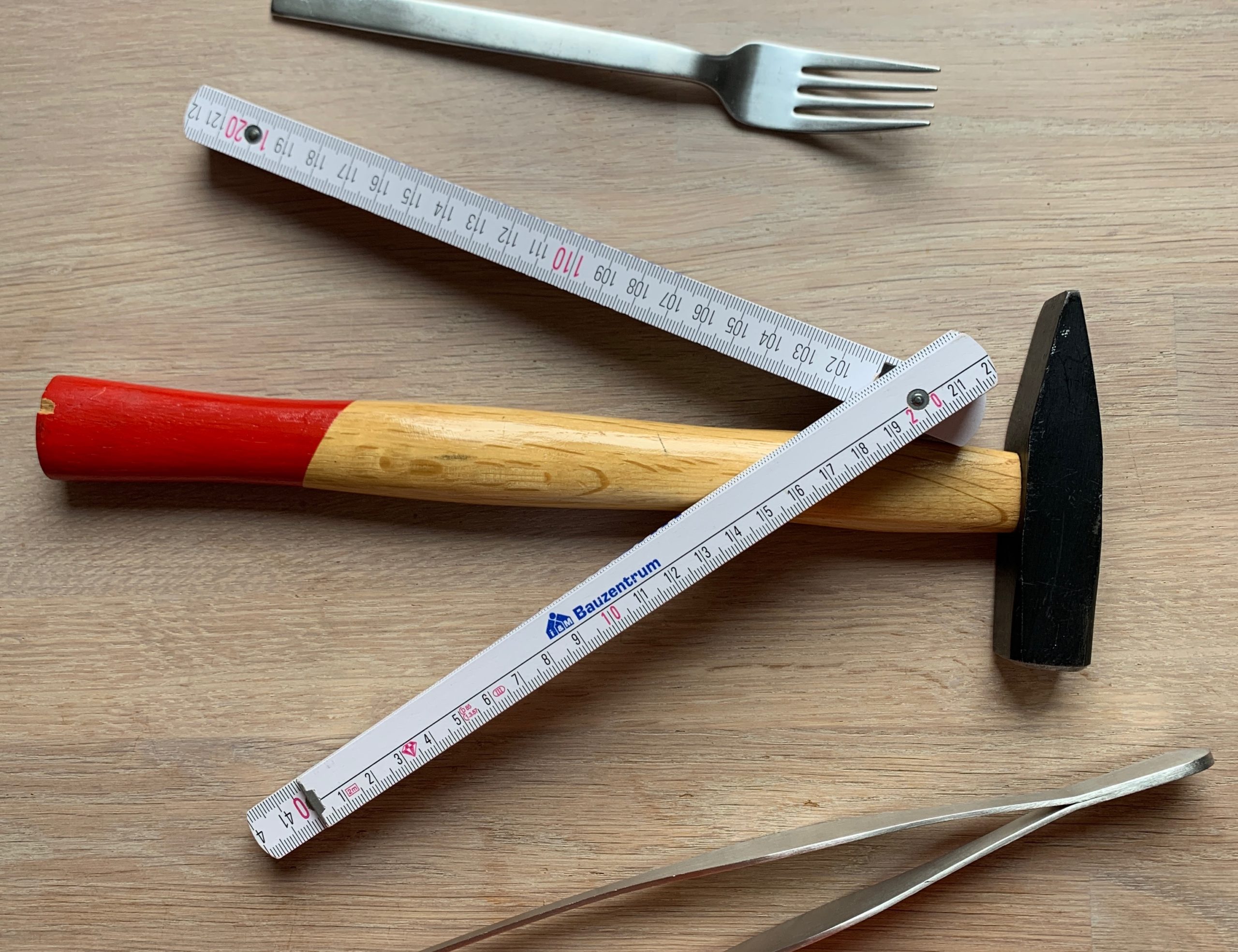 Ein Hammer, ein Zollstock, eine Gabel und eine Pinzette werden auf eine Holzfläche gelegt. Ein Hammer, ein Zollstock, eine Gabel und eine Pinzette werden auf eine Holzfläche gelegt.