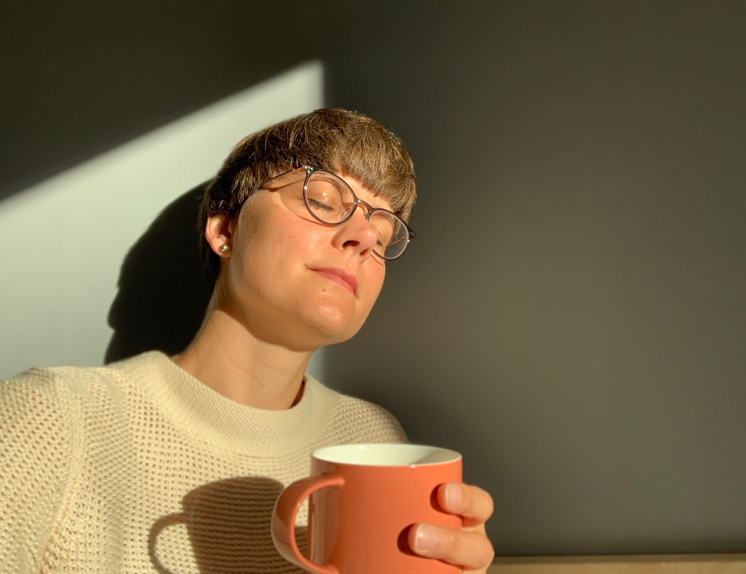 Eine Person mit Brille und cremefarbenem Pullover sitzt im Sonnenlicht, hält einen rosa Becher in der Hand und wirft einen Schatten an die Wand hinter sich.