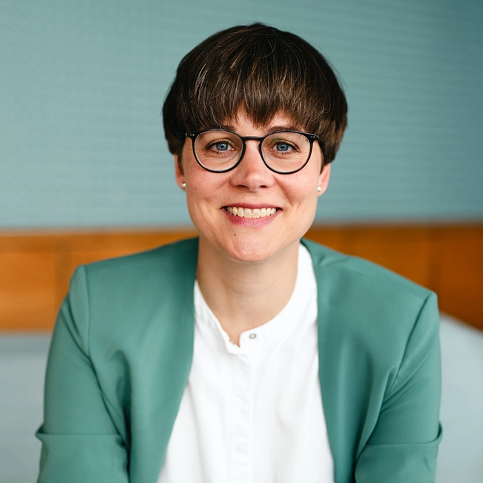 Sabrina Egly Sabrina Egly sitzt lächelnd in einem Innenraum vor einem türkisen und holzgetäfelten Hintergrund. Sie trägt eine Brille, ein weißes Hemd und einen grünen Blazer.