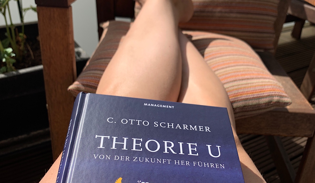 Auf einem Stuhl sitzende Person mit ausgestreckten Beinen, die das Buch "Theorie U" von C. Otto Scharmer hält. Im Hintergrund sind ein gestreiftes Kissen und ein Teil einer Pflanze zu sehen.