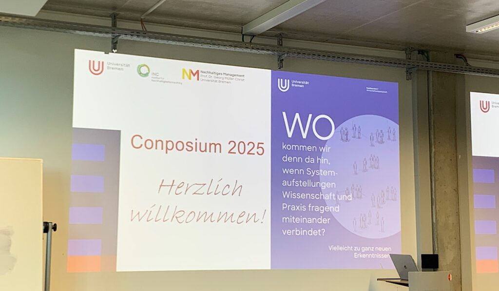 Auf einer großen Projektionsfläche wird "Conposium 2025. Herzlich willkommen!" und ein Veranstaltungsplakat mit deutschem Text zur Verbindung von Wissenschaft und Praxis in einem modernen Konferenzraum.