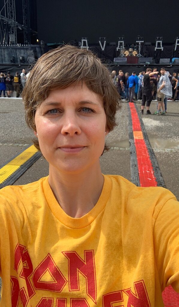 Eine Person, die ein gelbes Iron Maiden-Shirt trägt, macht ein Selfie bei einem Konzert im Freien mit einer Bühne und Menschen im Hintergrund.