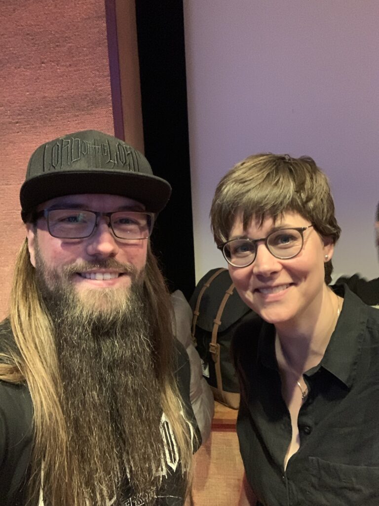 Zwei Personen lächeln für ein Selfie in einem Haus. Die Person auf der linken Seite hat lange Haare, einen dichten Bart, eine Brille und eine schwarze Mütze. Die Person auf der rechten Seite hat kurze Haare, eine Brille und trägt ein schwarzes Hemd. Im Hintergrund ist ein Rucksack zu sehen.