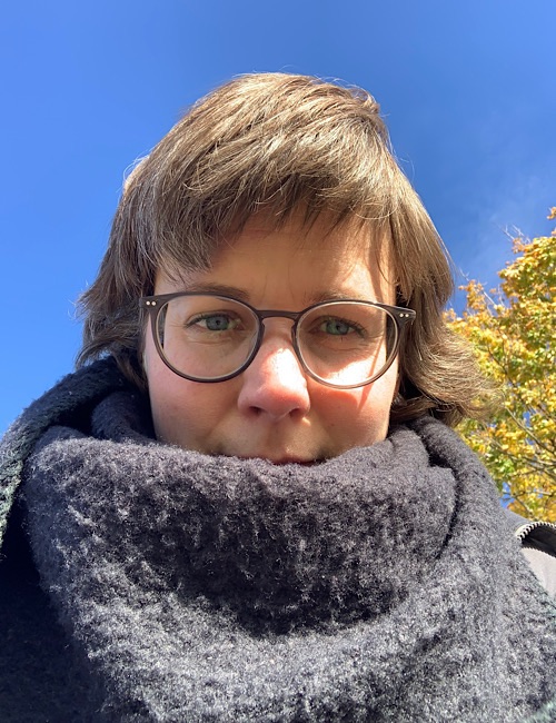 Eine Person mit kurzen braunen Haaren und Brille ist in einen dicken schwarzen Schal gehüllt und steht an einem sonnigen Tag im Freien, mit einem klaren blauen Himmel und Herbstblättern im Hintergrund.