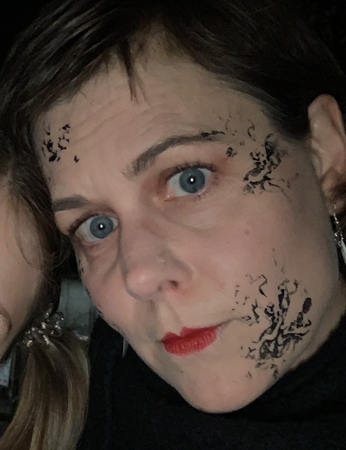 Eine Person mit kurzen braunen Haaren, blauen Augen, rotem Lippenstift und schwarzer abstrakter Gesichtsbemalung auf Stirn und Wange blickt direkt in die Kamera. Sie trägt ein schwarzes Oberteil und silberne Ohrringe.