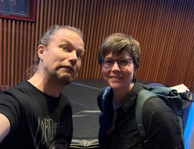 Zwei Personen stehen in einem Innenraum vor einer Holzlattenwand. Eine Person mit langen Haaren macht ein lustiges Gesicht, und die andere, die eine Brille und einen Rucksack trägt, lächelt in die Kamera. Die Beleuchtung ist warm und weich.