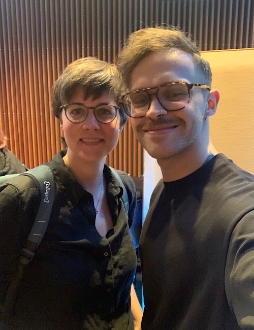 Zwei Personen mit Brillen und schwarzen Oberteilen lächeln für ein Selfie in Innenräumen in die Kamera. Einer hat kurze Haare und einen Rucksack, der andere hat gestylte Haare und Gesichtsbehaarung. Im Hintergrund sind vertikale Holzlatten zu sehen.