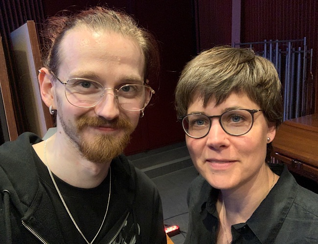 Zwei Personen mit Brillen stehen in einem Innenraum dicht beieinander, schauen in die Kamera und lächeln leicht. Im Hintergrund sind dunkle Holzpaneele und Metallrohre zu sehen.