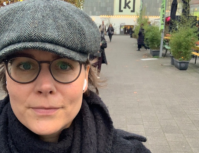 Eine Person, die eine Brille, eine flache Mütze und dunkle Kleidung trägt, macht ein Selfie im Freien auf einem gepflasterten Gehweg, wobei im Hintergrund Menschen und Grünflächen zu sehen sind.