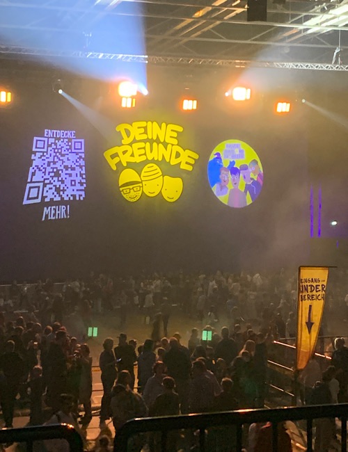 Eine Menschenmenge versammelt sich in einer Konzerthalle mit Scheinwerfern und projizierten Grafiken an der Wand, darunter der Text "DEINE FREUNDE" und Cartoon-Gesichter. Ein QR-Code und zusätzlicher deutscher Text werden ebenfalls angezeigt.