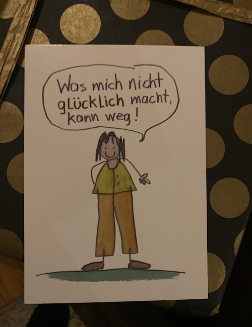 Eine handgezeichnete Illustration einer lächelnden Person mit braunem Haar, die eine grüne Weste und eine braune Hose trägt, sagt in einer Sprechblase: "Was mich nicht glücklich macht, kann weg!" (Was mich nicht glücklich macht, kann weg!).