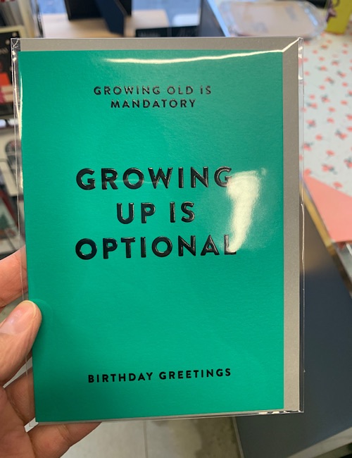 Eine Hand hält eine blaugrüne Geburtstagskarte mit schwarzer Schrift, auf der steht: "Alt werden ist Pflicht. Erwachsenwerden ist optional. Geburtstagsgrüße." Die Karte befindet sich in einer Plastikhülle.