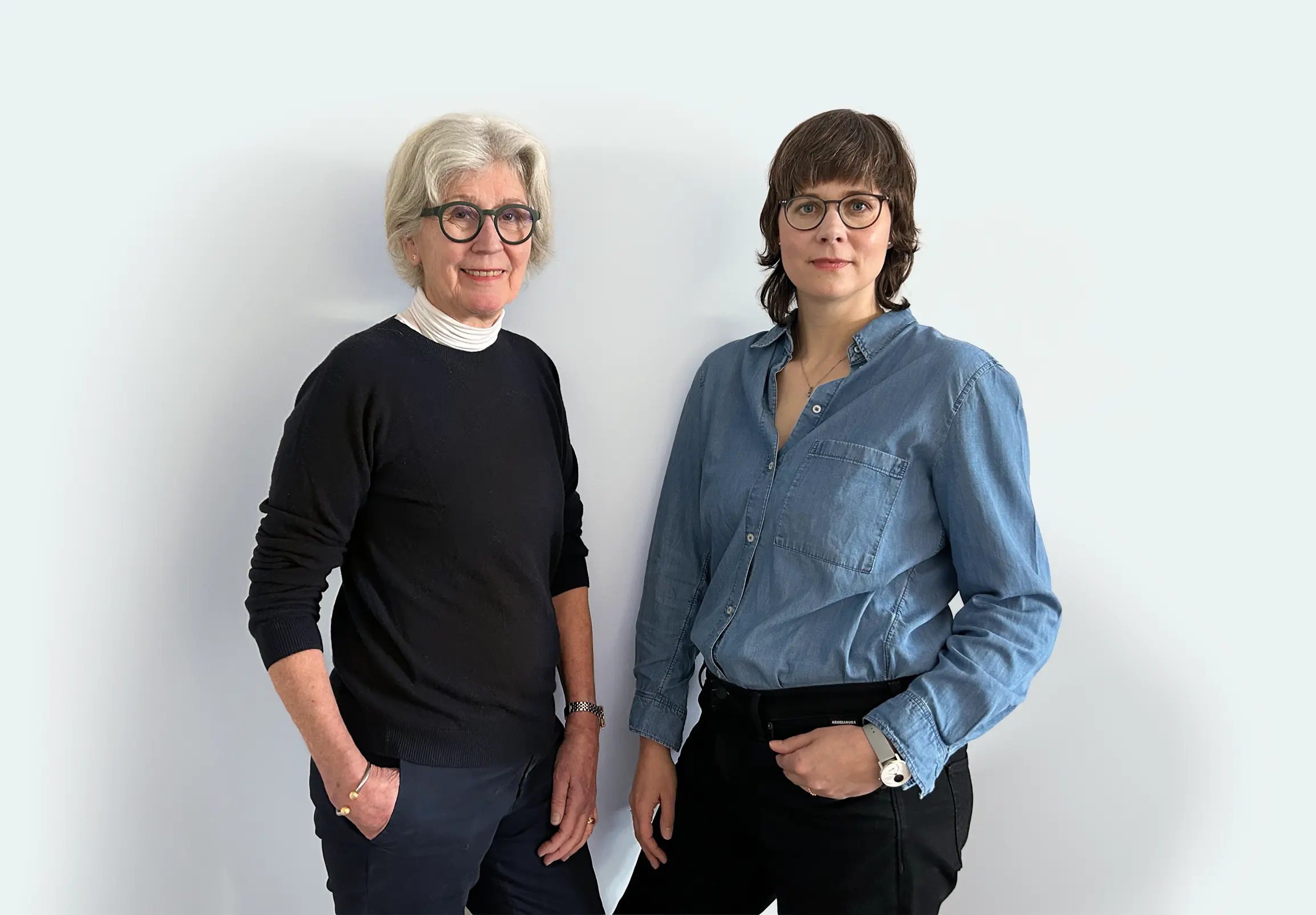 Andrea und Sabrina stehen nebeneinander vor einem schlichten hellen Hintergrund, beide tragen eine Brille. Andrea hat graues Haar und trägt einen dunklen Pullover über einem weißen Oberteil, Sabrina hat kurzes braunes Haar und trägt ein blaues Button-up-Hemd. Beide haben die Hände in den Taschen.