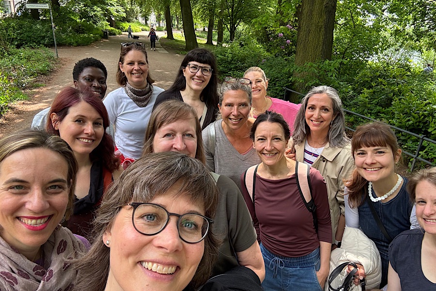 Eine Gruppe von zwölf lächelnden Frauen macht an einem hellen Tag in einem grünen, begrünten Park ein gemeinsames Selfie. Einige tragen Brillen, und alle wirken glücklich und entspannt.