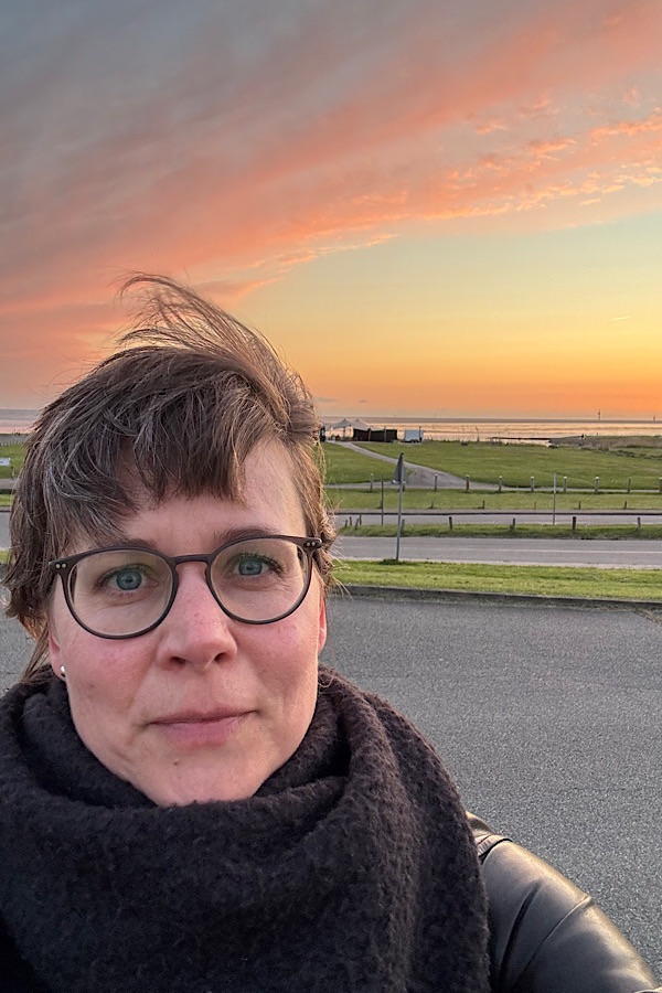 Eine Person mit kurzen Haaren und Brille, die einen schwarzen Schal trägt, steht im Freien, mit einem malerischen Sonnenuntergangshimmel, grasbewachsenen Feldern und einem Gewässer im Hintergrund.