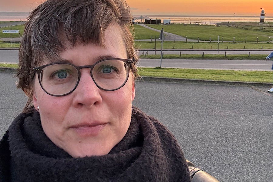 Eine Frau mit kurzen braunen Haaren, einer Brille und einem schwarzen Schal macht ein Selfie im Freien bei Sonnenuntergang. Hinter ihr sind eine Straße, eine Wiese, Wasser und ein Leuchtturm am Horizont zu sehen.