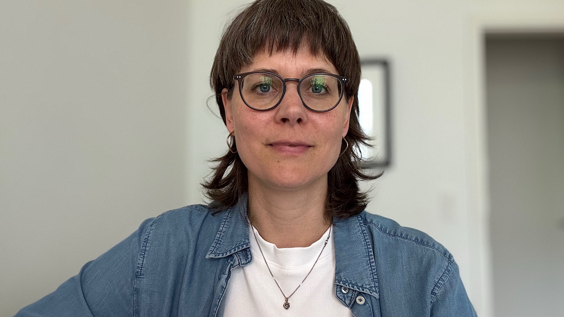 img_2184 Eine Person mit kurzen braunen Haaren und Brille, die ein Jeanshemd über einem weißen T-Shirt und zwei Halsketten trägt, blickt in einem hellen Raum mit weißen Wänden in die Kamera.